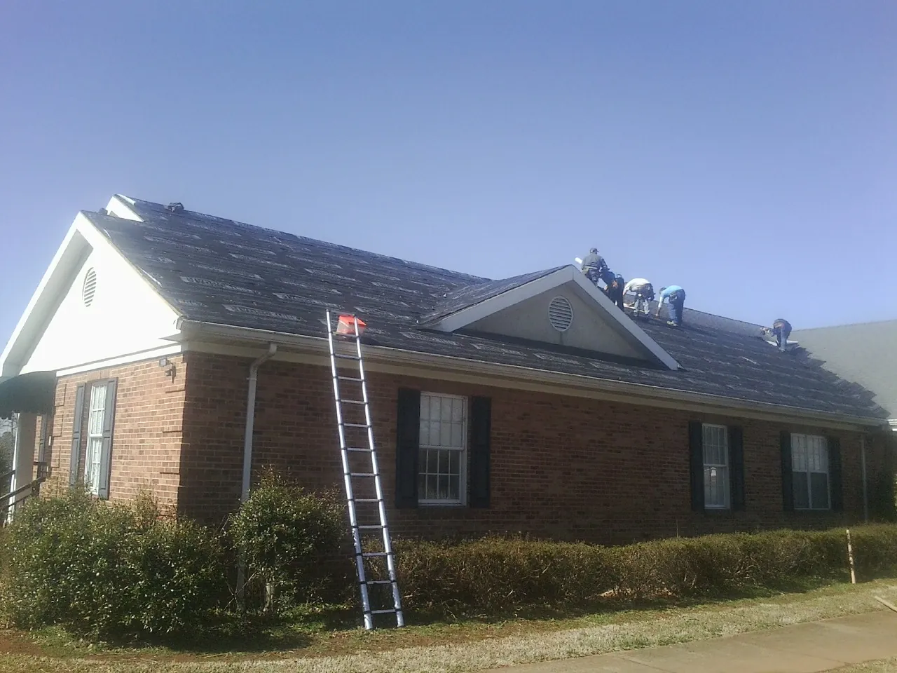 Crew tearing shingles off a brick ranch roof with a ladder set — Emergency Roof Repair work by our crew in Elmsford