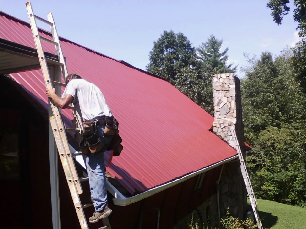 Roofer on a ladder beside a red metal roof and stone chimney — Soffit & Fascia Repair by Roofing Brothers in Elmsford